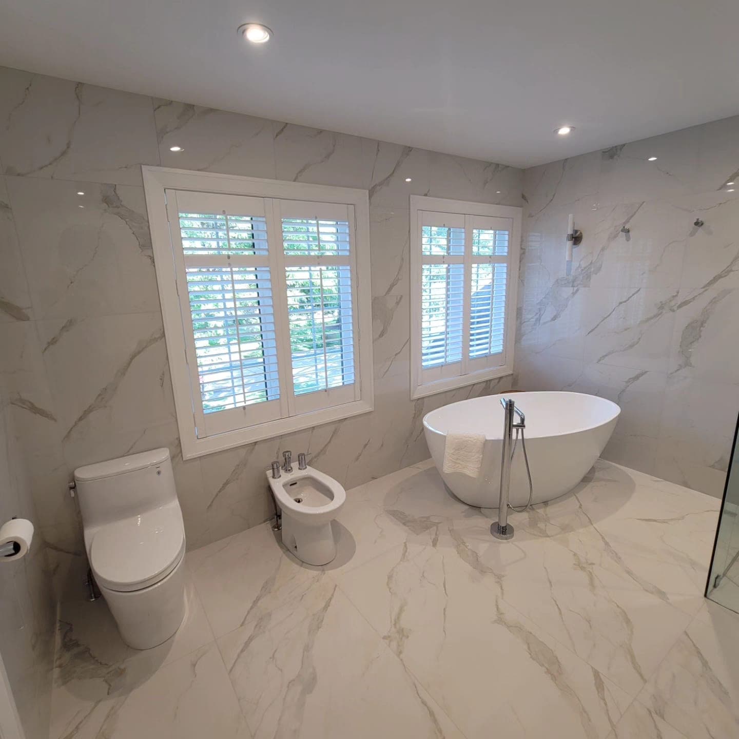 Freestanding tub against full-height Calacatta marble wall with plantation-shutter window light
