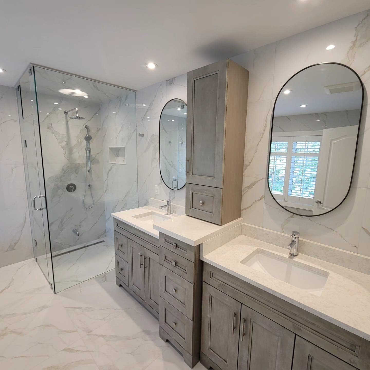 Double vanity with grey shaker cabinets, oval mirrors, and walk-in glass shower against Calacatta marble walls